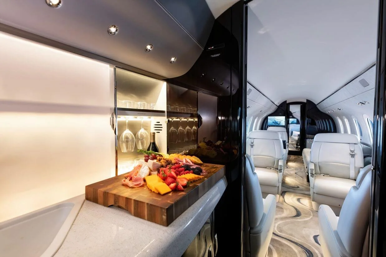 Cessna Citation Longitude  Luxury private jet interior with modern seats, ambient lighting, and a galley featuring a wooden board with assorted fruits, cheese, and cured meats.