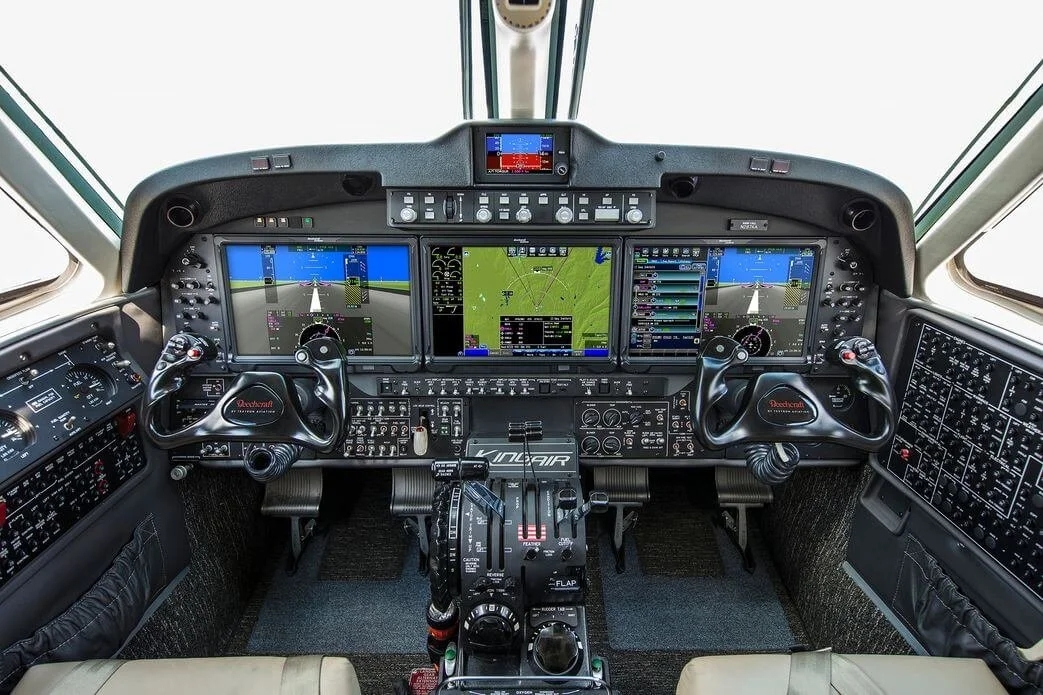 Cockpit of Beechcraft King Air 350 Charter featuring multiple flight controls, digital displays, and instrument panels. Two yokes are present with various switches and knobs, alongside navigation and communication equipment.