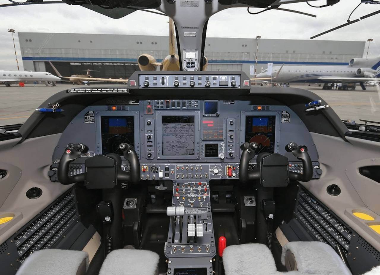Private jet cockpit with advanced avionics displays and controls, parked on an airfield with jet aircraft in the background.