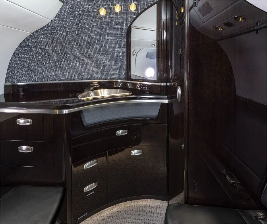 Interior view of an airplane lavatory with dark wood finishes, a countertop with a metallic sink, storage drawers, and textured wall design.