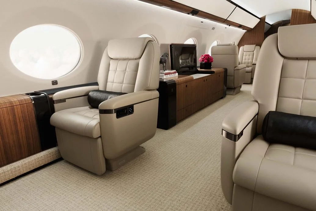 Luxury private jet interior with beige leather seats, wood paneling, windows, and a central cabinet with flowers and decor.