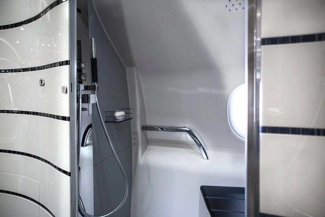 Interior of an aircraft bathroom with a shower and curved design, featuring a showerhead, grab bar, and small shelf.