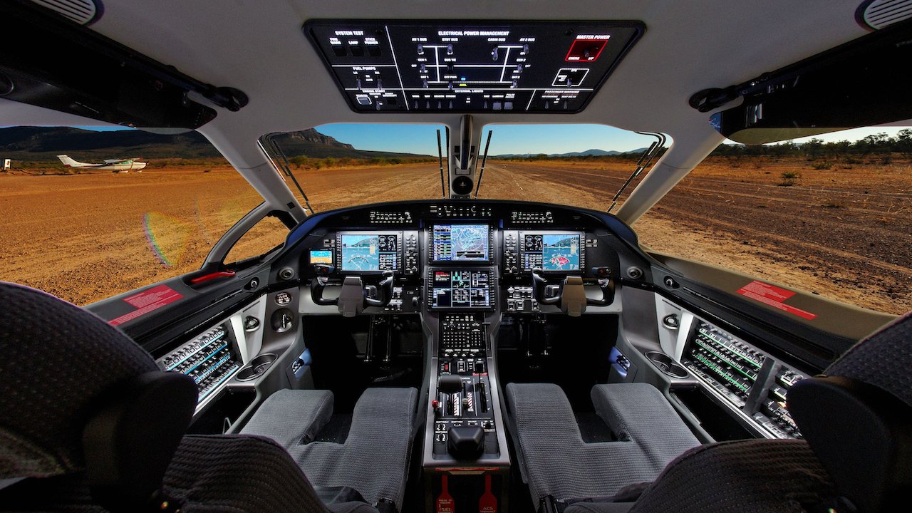 Cockpit of a modern aircraft with digital displays and control panels, showing an exterior view of a desert landscape with a small plane visible.