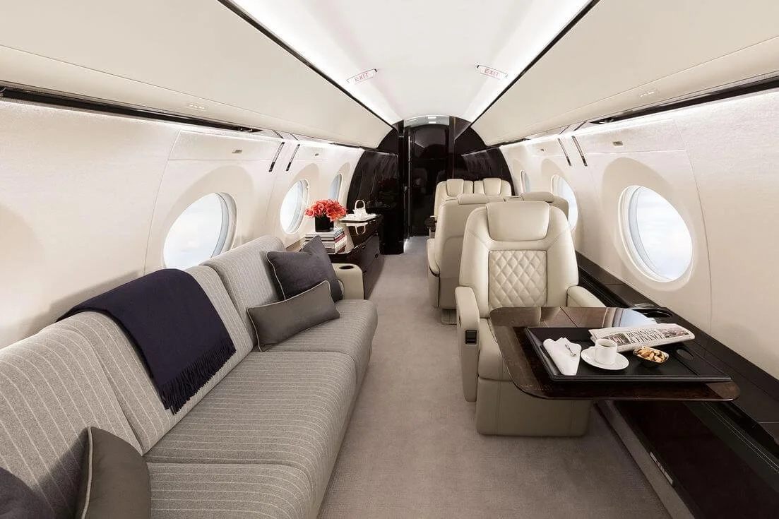 Gulfstream G500 Private Jet interior with beige leather seats, a gray sofa with cushions, a table set with a cup and saucer, and windows on the side.