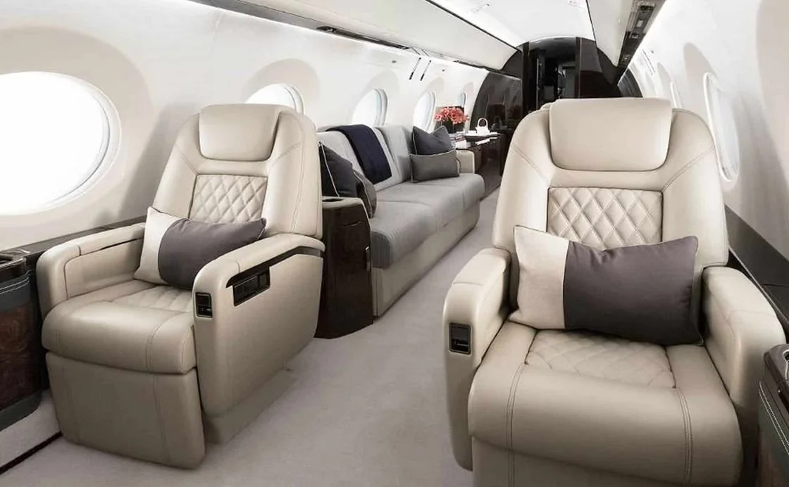 Luxury private jet interior with beige leather seats, plush cushions, and large windows.