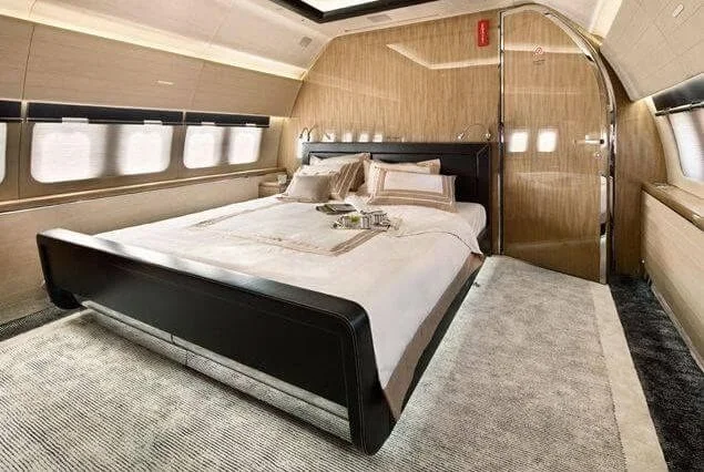 Luxurious private jet interior with a king-size bed, beige bedding, and elegant wooden paneling.