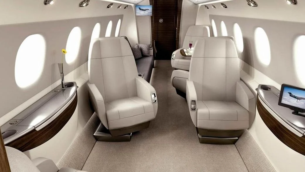 Interior of a luxury private jet Embraer Legacy 450 with leather seats, plush carpeting, and small windows. There is a flower on a side table and a small screen displaying an image of an aircraft. The aisle leads to the back with additional seating a