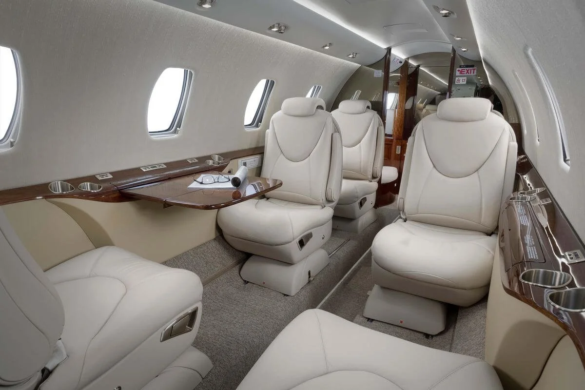 Interior of a private jet with beige leather seating, a fold-out table, windows, and overhead lighting.