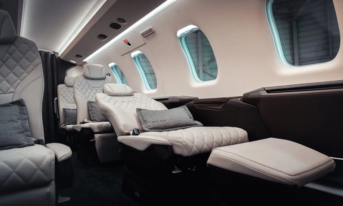  interior OF Pilatus PC24 Private Jet Charter  with plush white leather seats, quilted design, and matching gray cushions. Three windows line the wall, with soft lighting creating an elegant atmosphere.