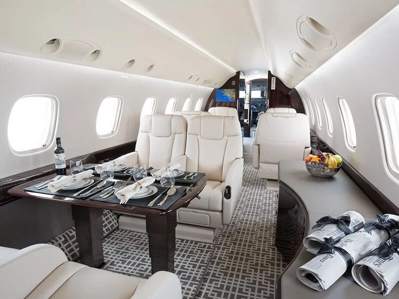 Luxury private jet interior with cream leather seats, dining table set with wine and glasses, fruit bowl on side table, and rolled newspapers.