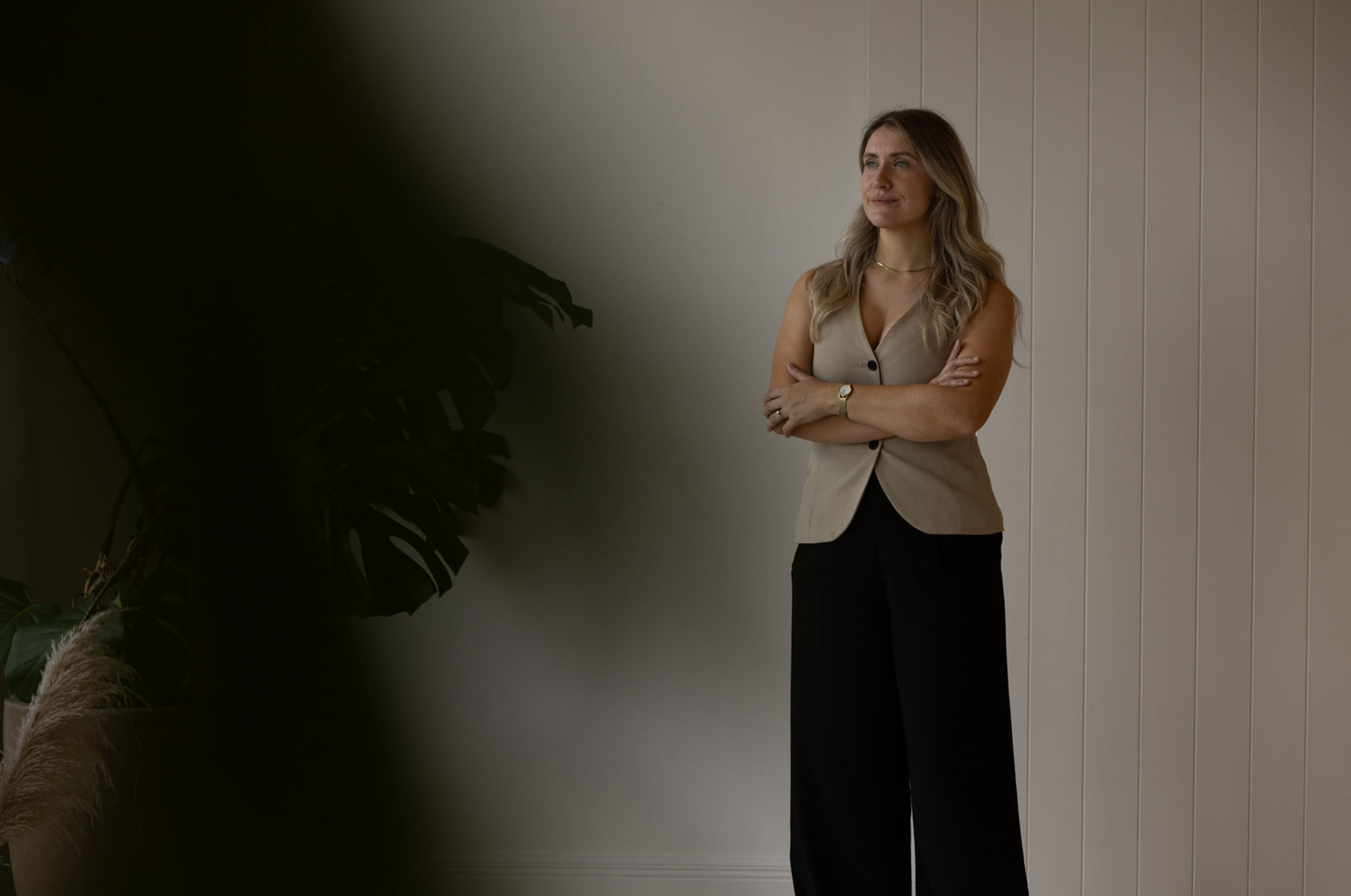 A woman with light brown, wavy hair standing against a white paneled wall, crossing her arms, wearing a beige sleeveless vest, black wide-leg pants, a watch, and a gold necklace.