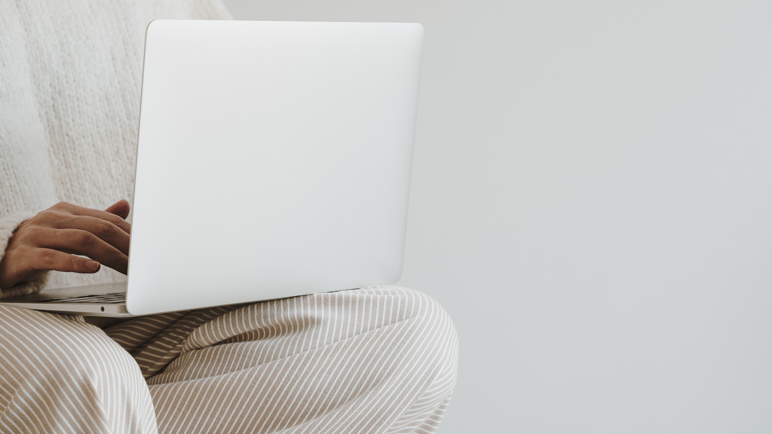 Person sitting on a beige and white striped lounge chair using a silver laptop, with a plain white wall in the background.