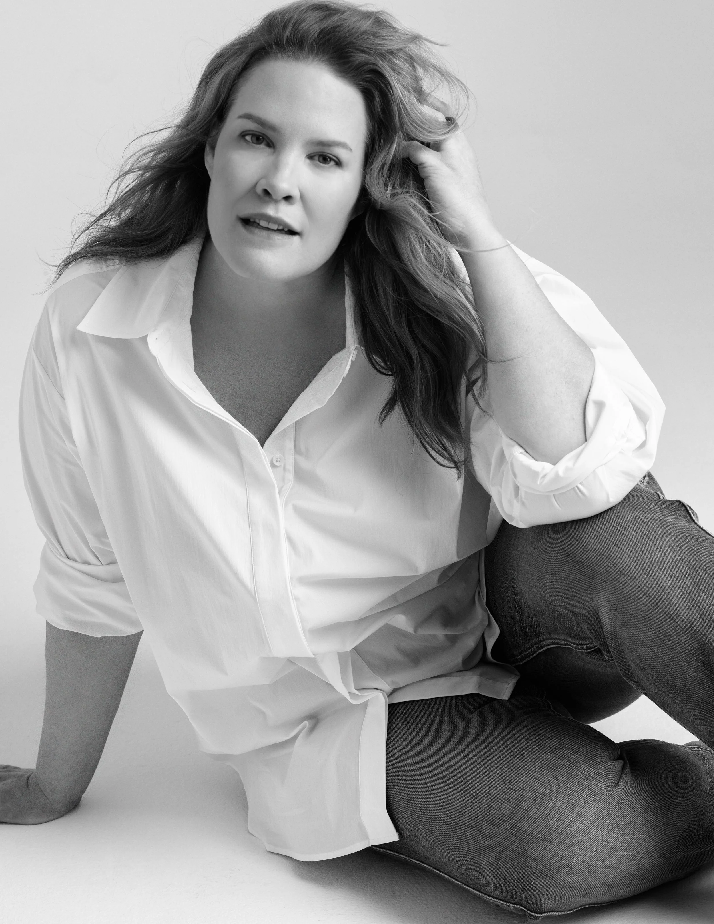 Black and white photo of a woman with long, wavy hair, sitting on the floor with her legs crossed, wearing a white shirt and dark jeans, looking at the camera with one hand in her hair.