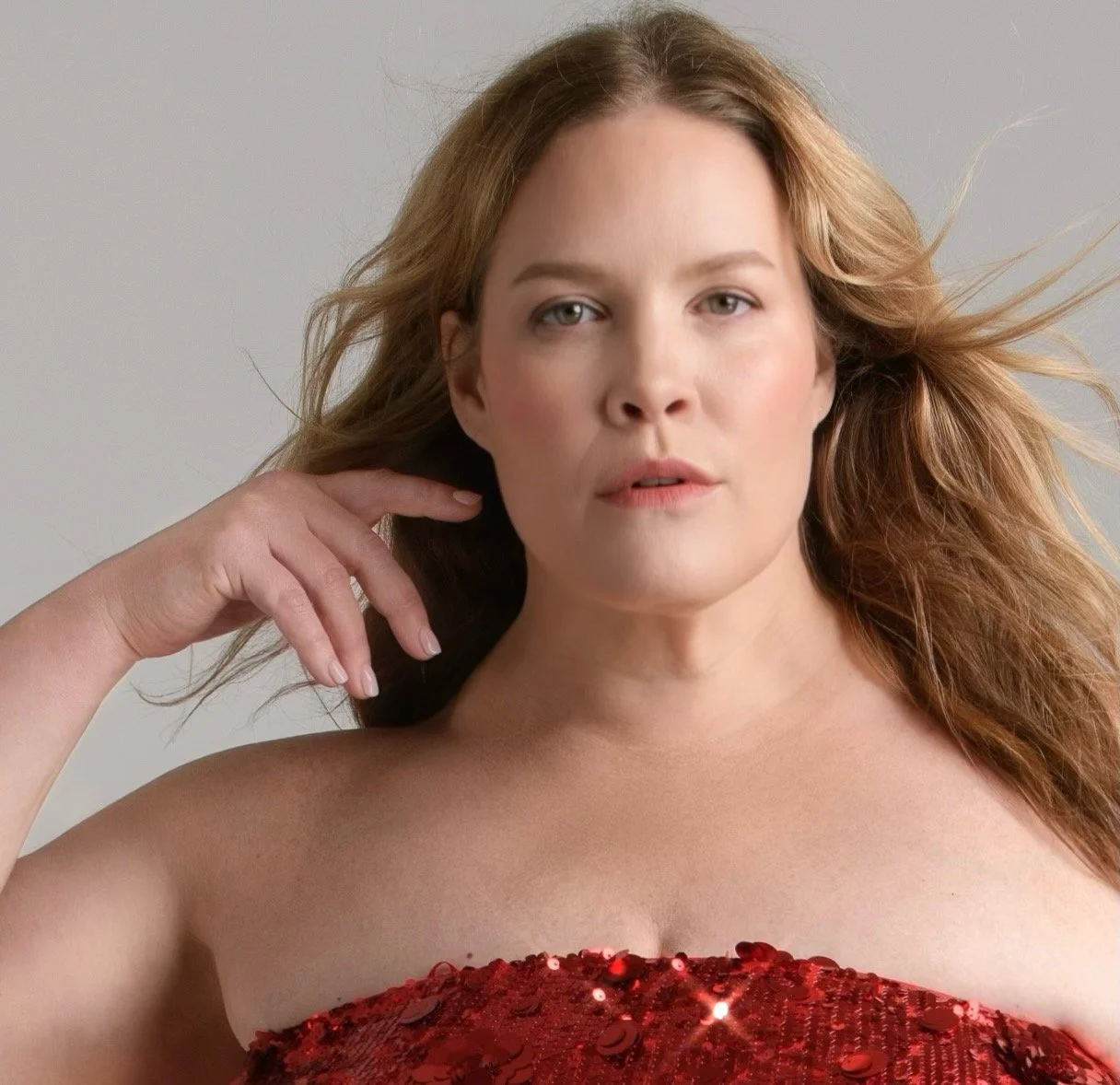 Close-up of a woman with wavy, light brown hair, wearing a strapless red dress with sequins, looking at the camera with a neutral expression.