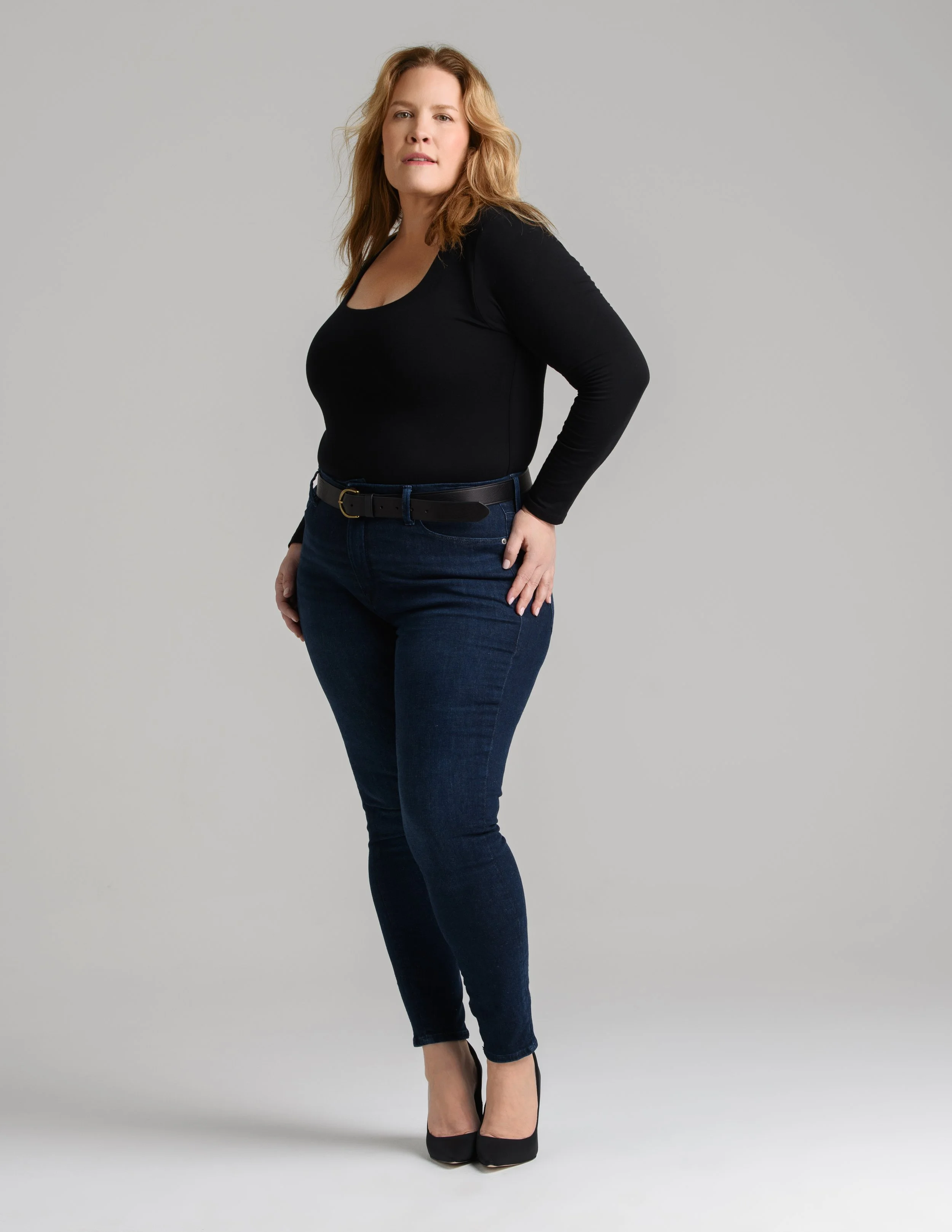 A woman standing against a plain gray background, wearing a black long-sleeve top, dark blue jeans, and black high heels.