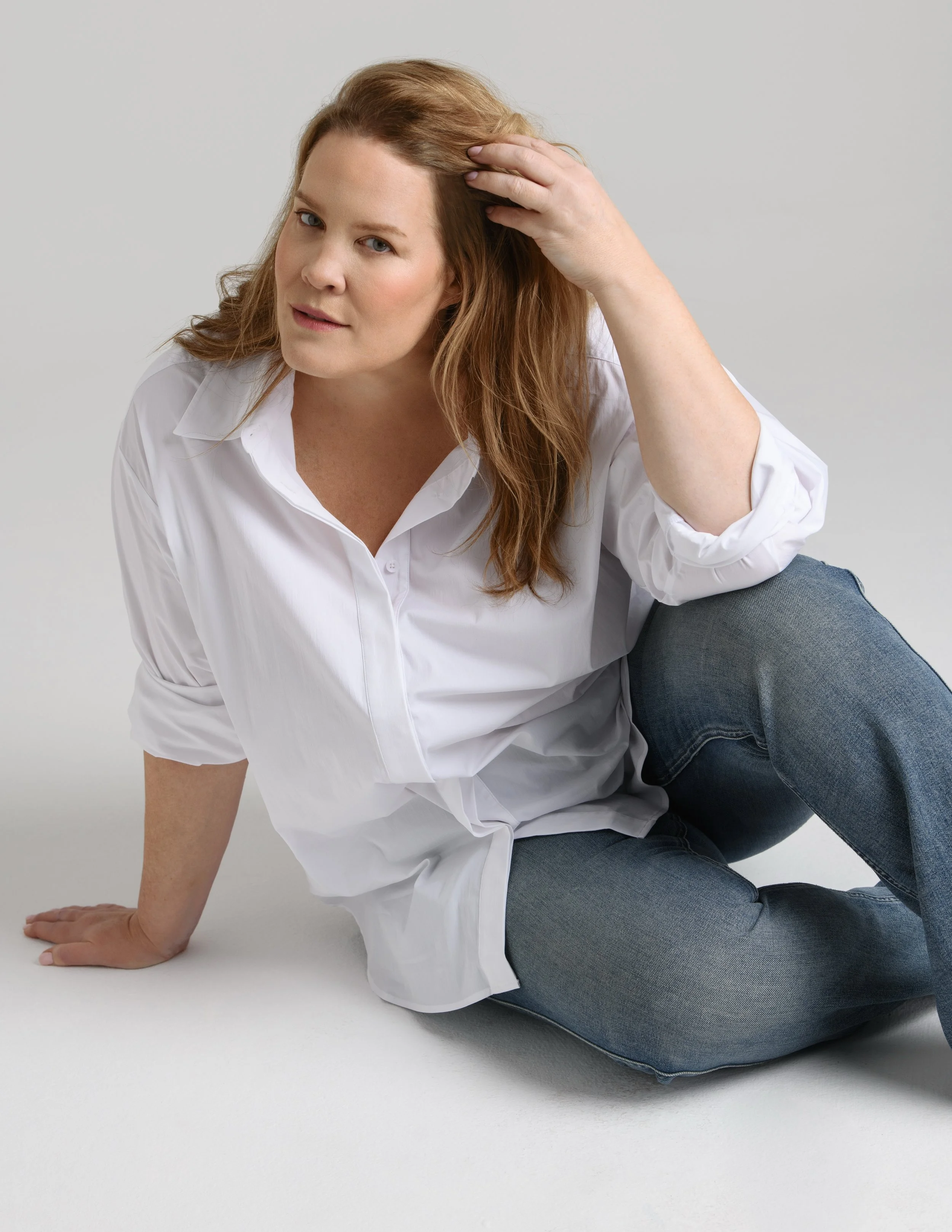 A woman with red hair and fair skin sitting on the floor, wearing a white shirt and blue jeans, posed against a plain light gray background.