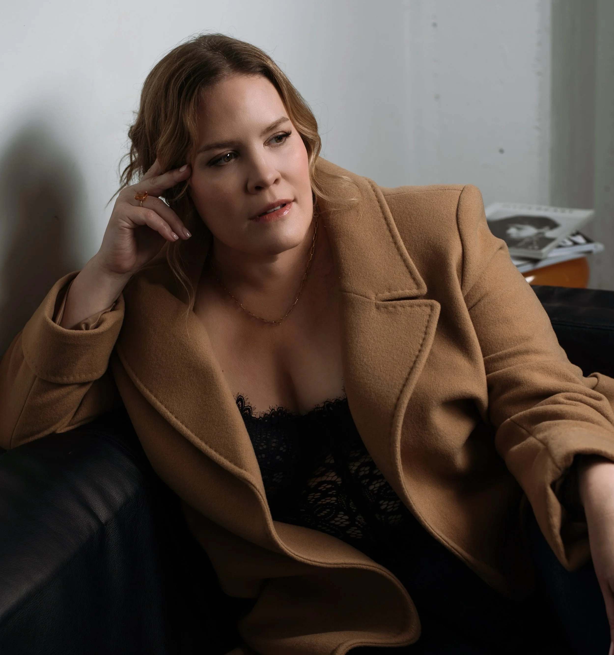 A woman with wavy, shoulder-length hair, wearing a tan coat and a black lace top, is sitting on a black leather couch with her head resting on her hand, looking thoughtful.
