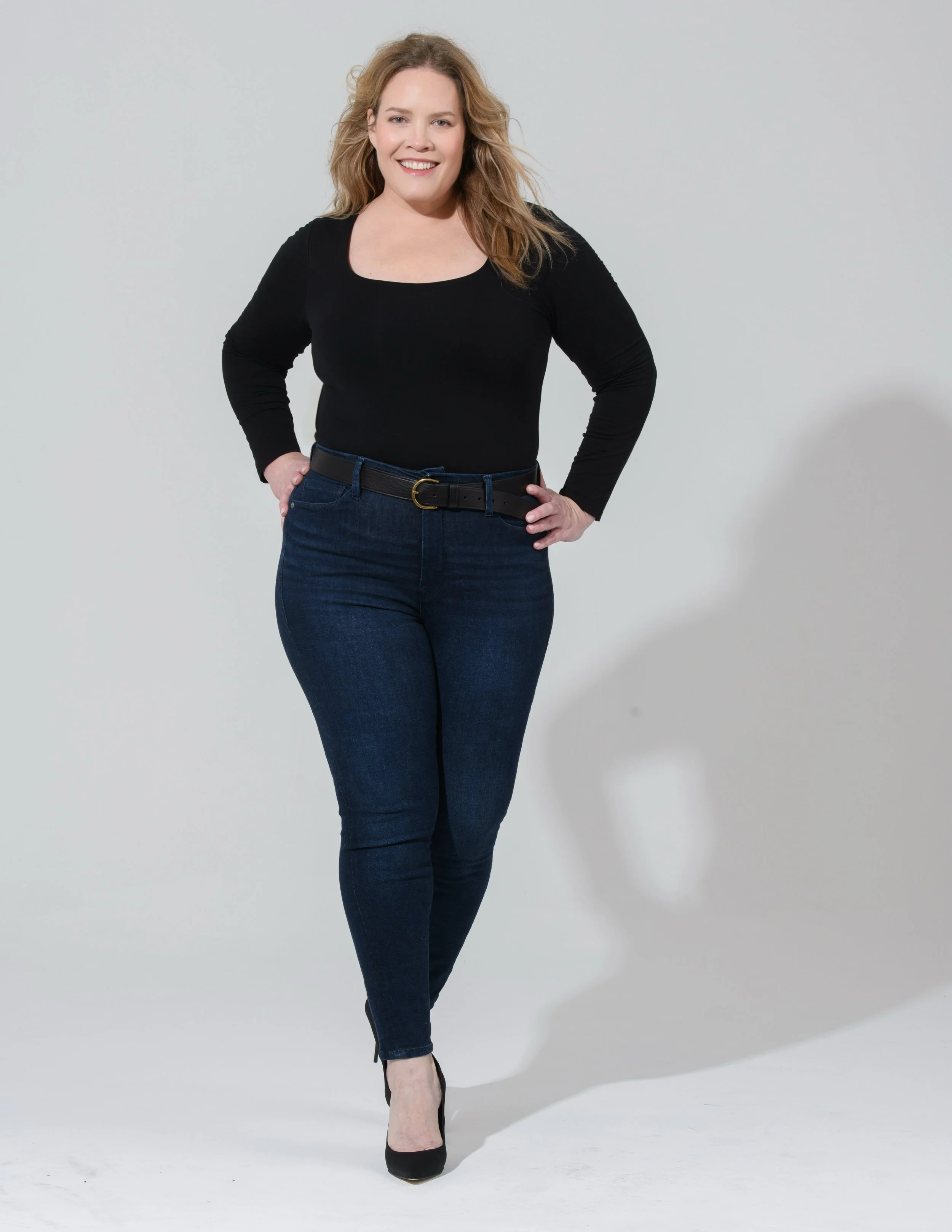 A smiling woman with blonde hair, dressed in a black top, dark blue jeans, and black heels, standing in front of a plain white background.