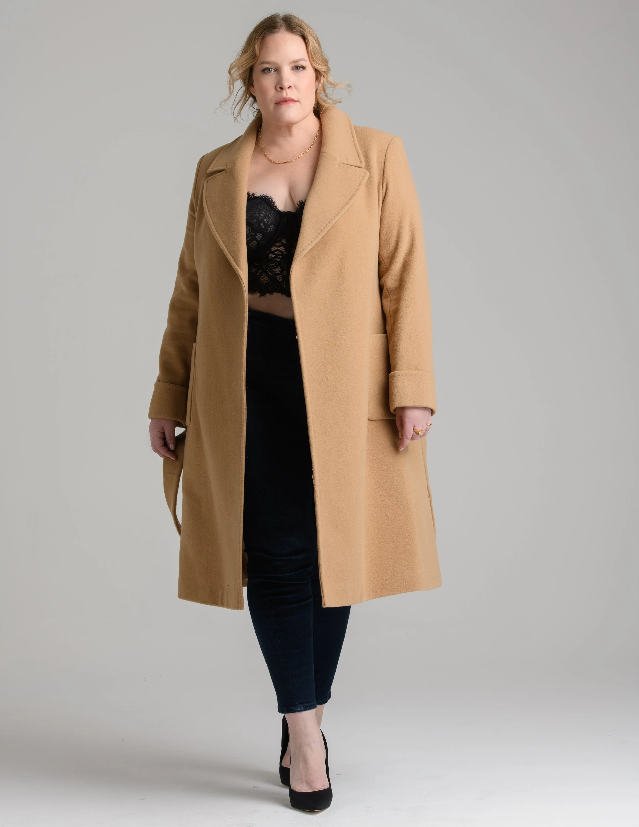 A woman wearing a tan coat, black lace top, black jeans, and black heels, standing against a plain background.