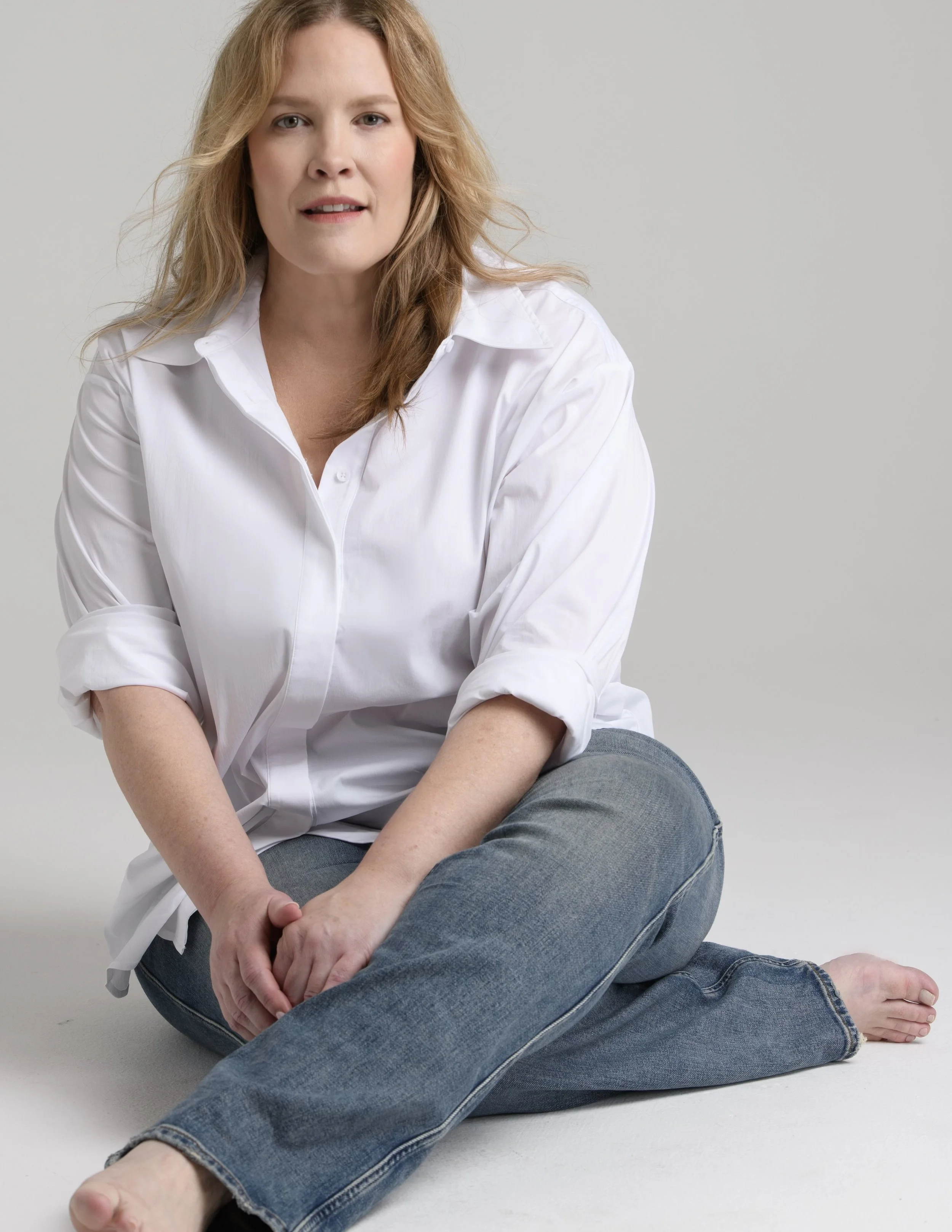A woman with red hair, wearing a white button-up shirt and blue jeans, sits on the floor with her legs crossed and arms resting on her knees, against a plain light gray background.