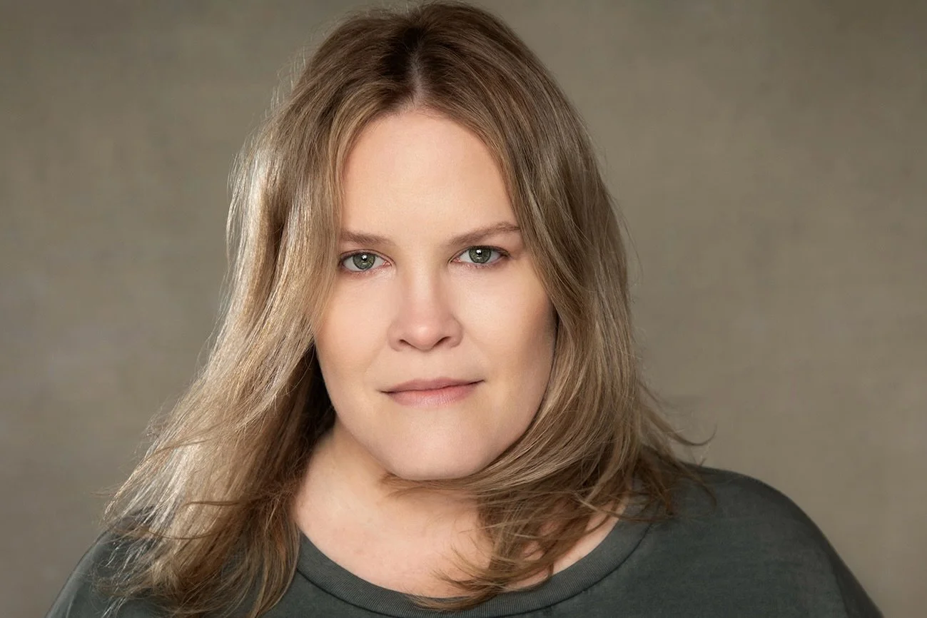 Close-up portrait of a woman with shoulder-length light brown hair and green eyes, wearing a dark shirt, against a neutral background.