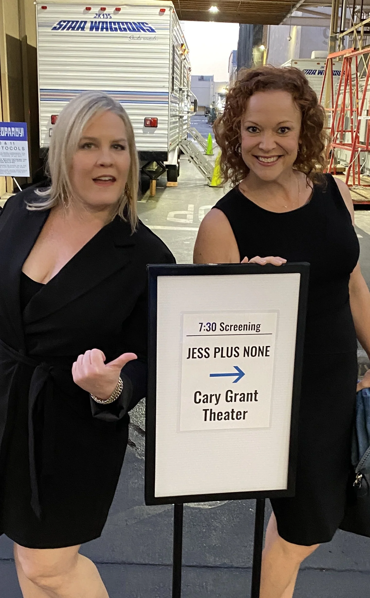 Two women standing next to a sign at a theater entrance, pointing towards the Cary Grant Theater. The sign indicates a 7:30 screening of 'Jess Plus None' and the women are smiling.