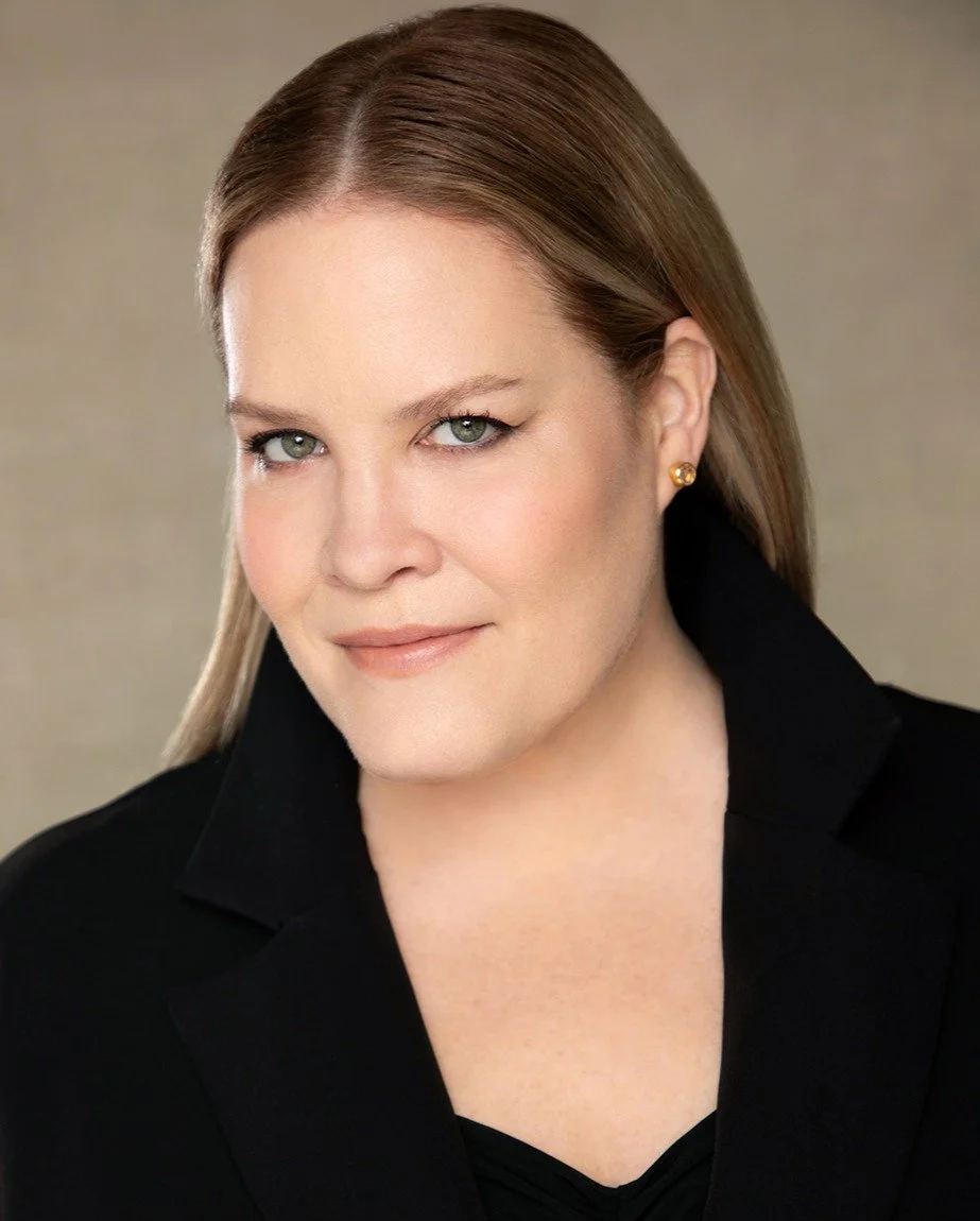 Close-up portrait of a woman with fair skin, straight light brown hair, blue eyes, and a slight smile, wearing a black blazer and gold earrings.