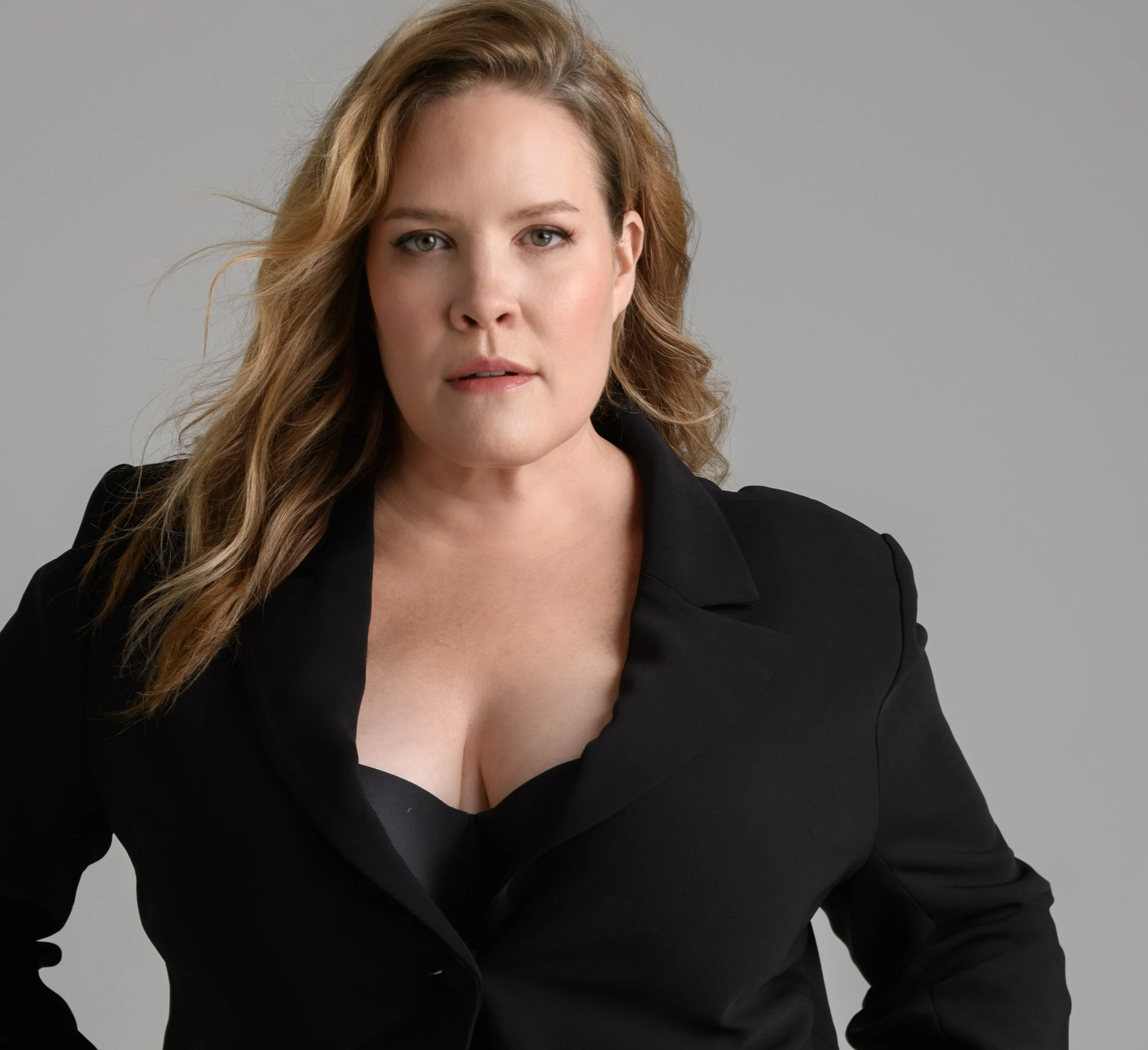 A woman with wavy blonde hair wearing a black blazer and black top against a neutral gray background.