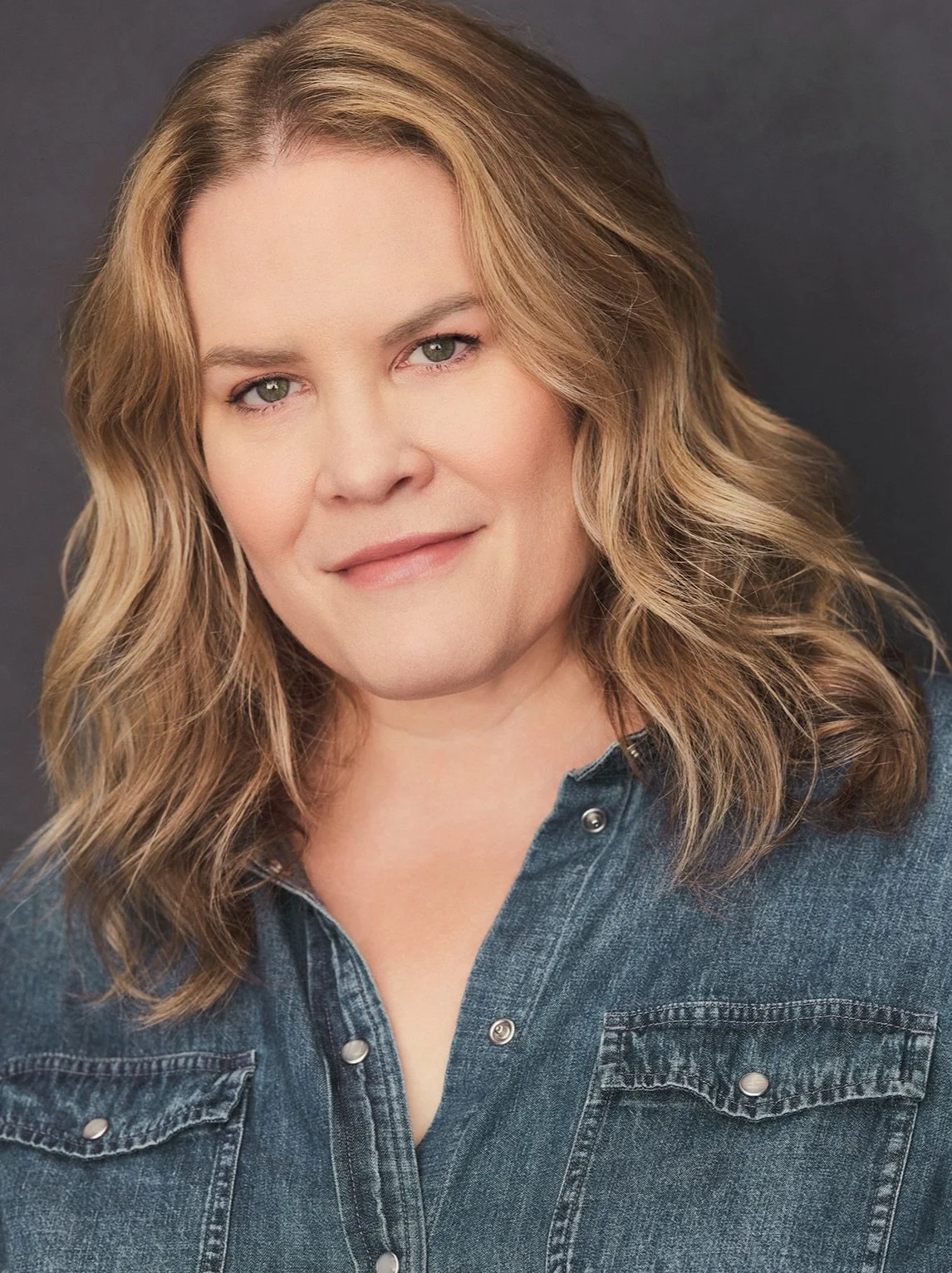 Close-up portrait of a woman with shoulder-length blond wavy hair, blue eyes, and fair skin, wearing a denim shirt with a collar and metallic buttons, smiling softly against a dark background.
