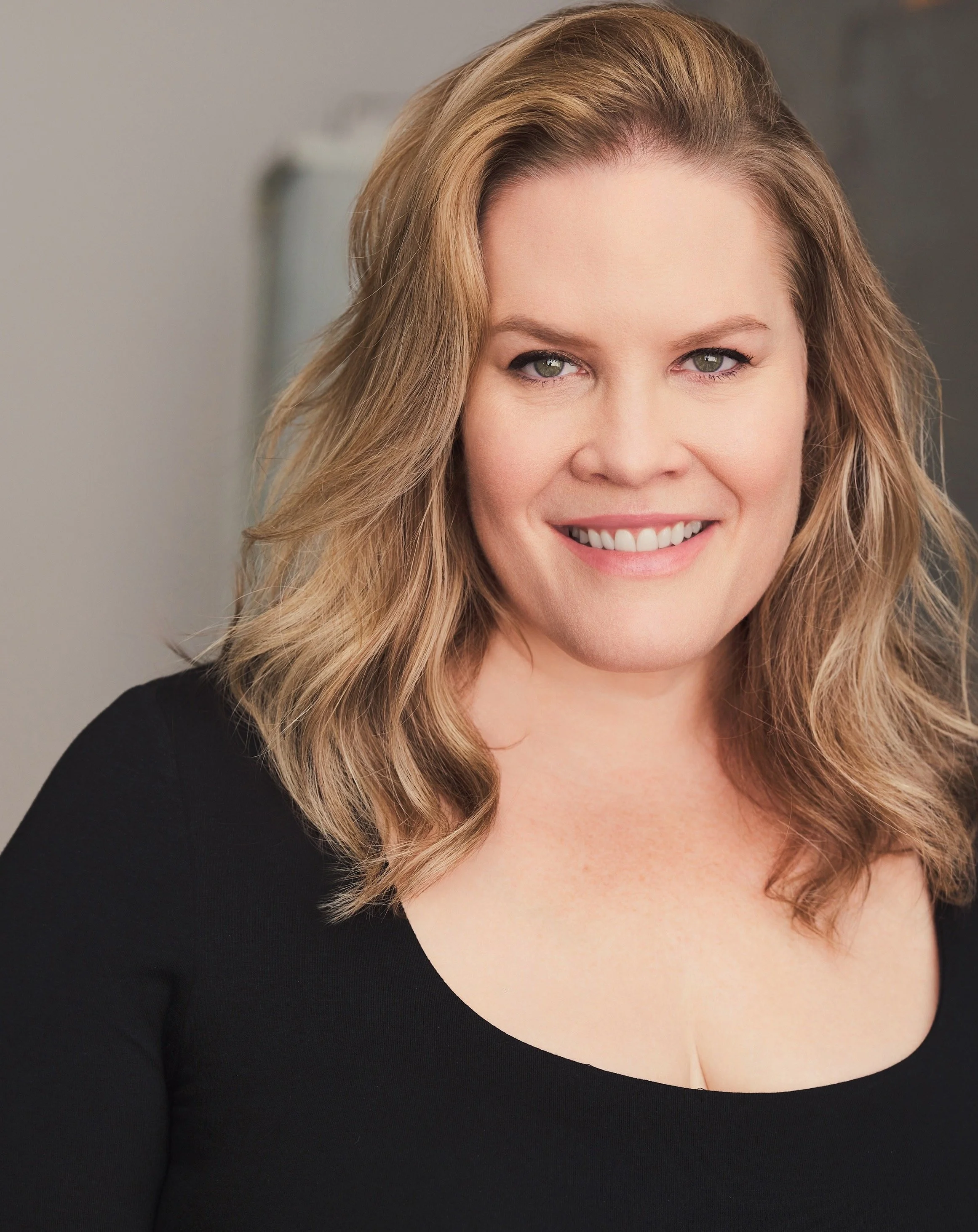 A woman with wavy blonde hair, blue eyes, and fair skin, smiling at the camera, wearing a black top.