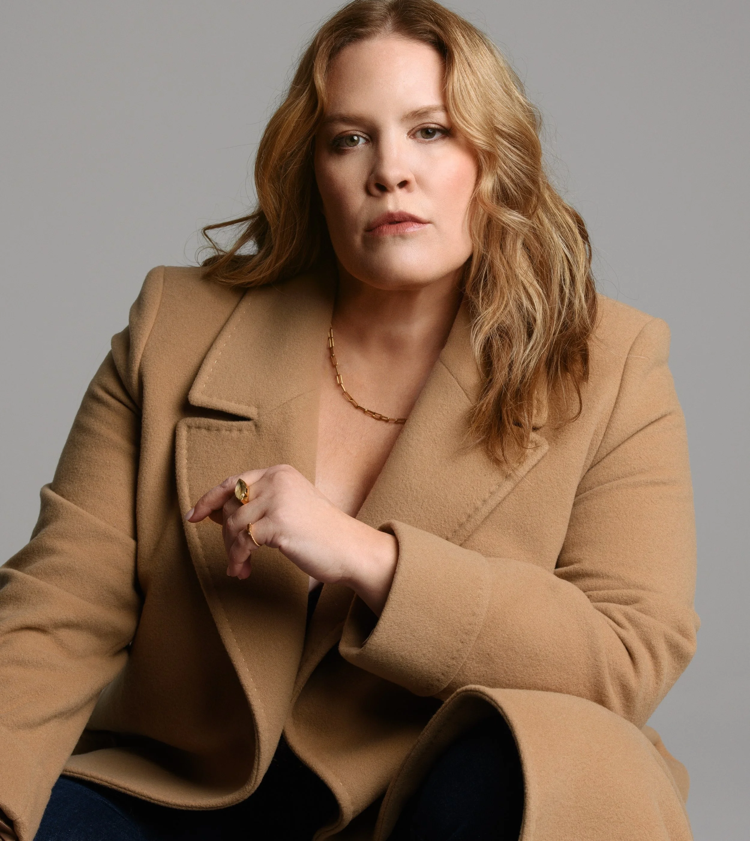 A woman with light skin, wavy, shoulder-length blonde hair, wearing a beige blazer and gold jewelry, sitting against a gray background.