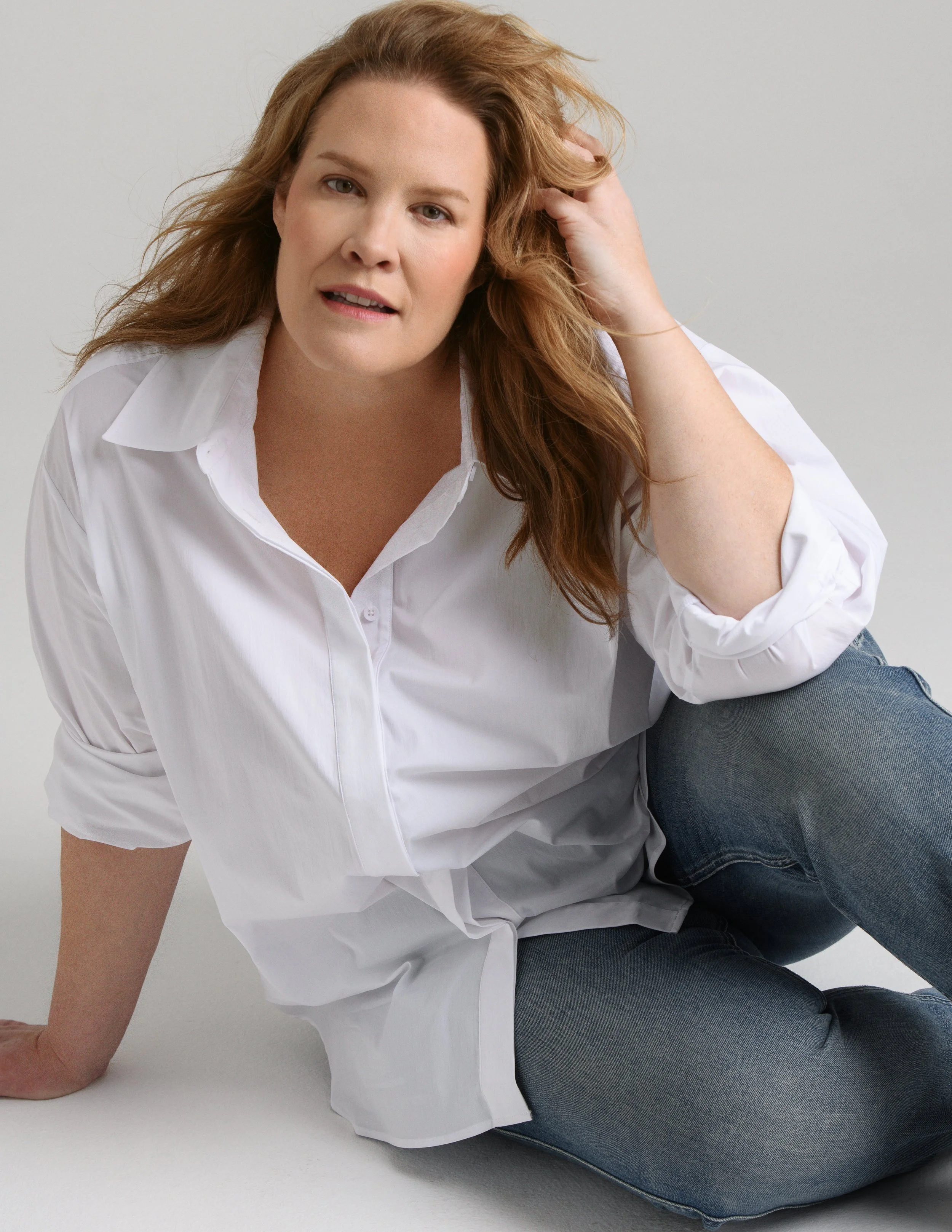 A woman with red hair wearing a white shirt and blue jeans, sitting on the floor.