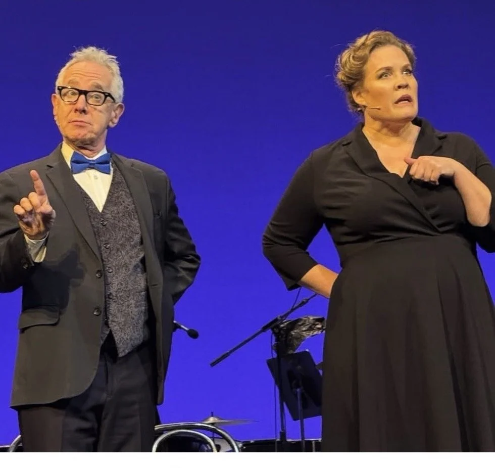 Two performers on stage, a man dressed in a tuxedo with a bow tie and glasses, and a woman in a black dress with a headset microphone. The man is holding up one finger and has a surprised expression, while the woman looks off to the side with an expressive face.