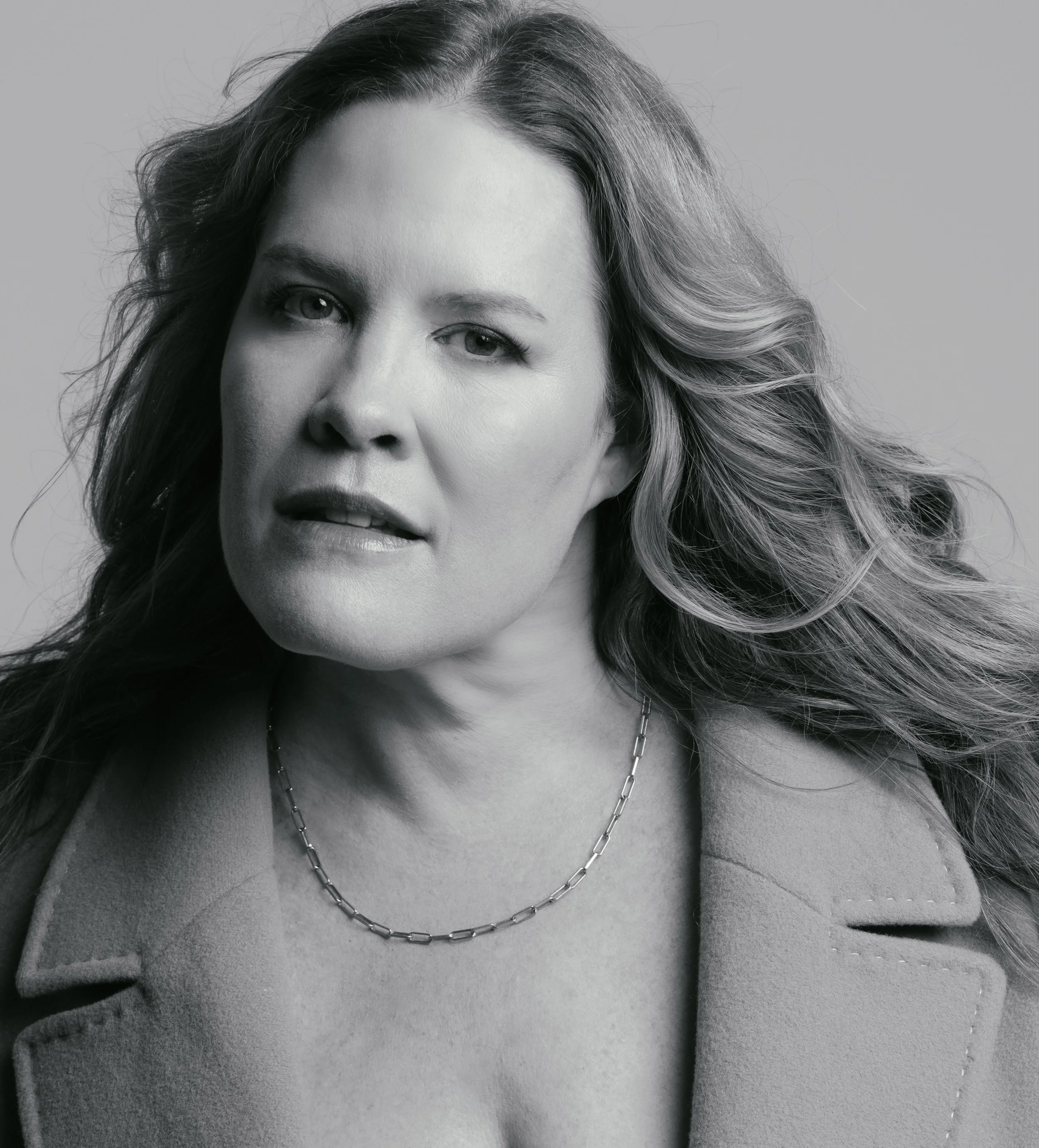 Black and white portrait of a woman with long wavy hair, wearing a coat and a chain necklace, looking directly at the camera.