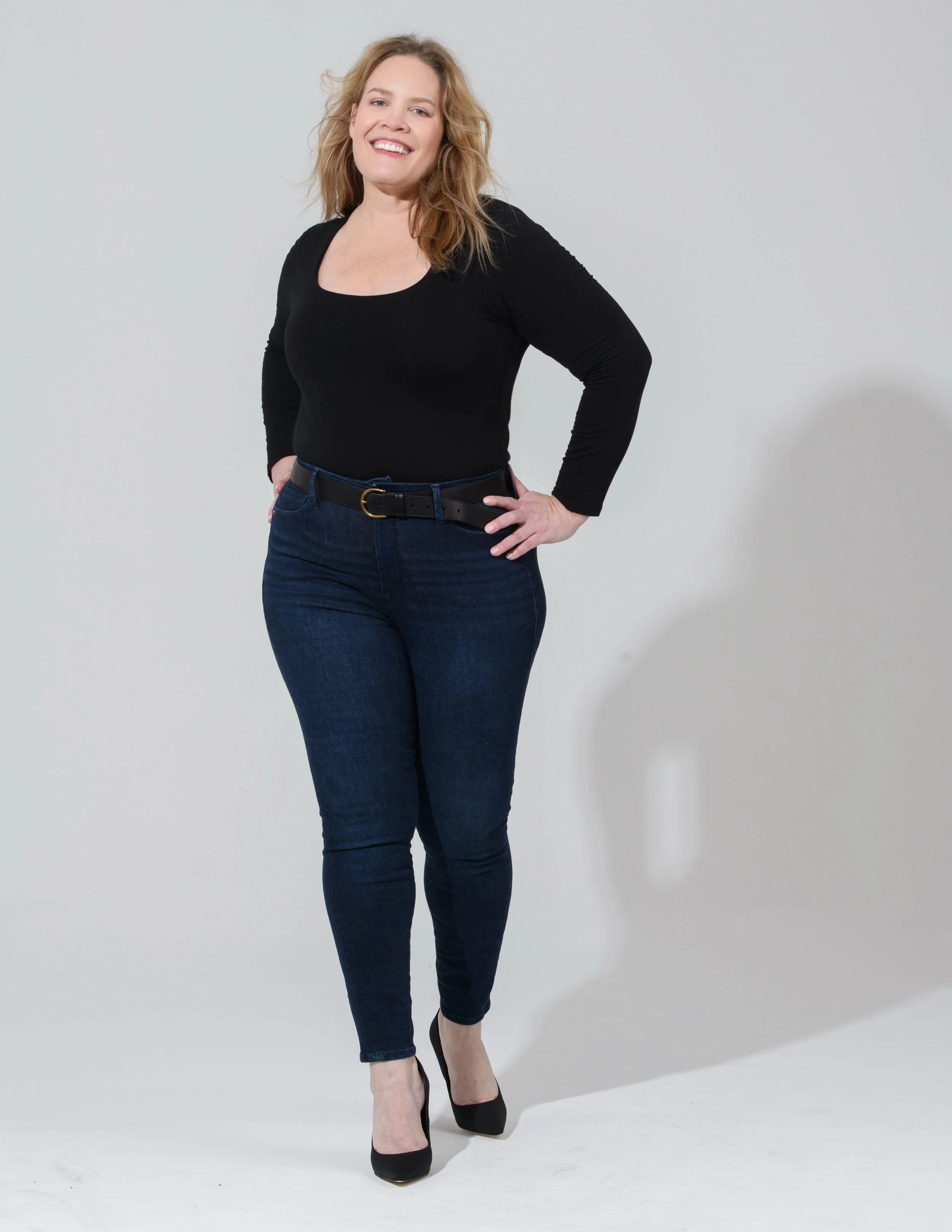 A woman with shoulder-length wavy hair, wearing a black long-sleeve top, dark blue jeans, black belt, and black heels, standing against a plain light-colored background, smiling with one hand on her hip.