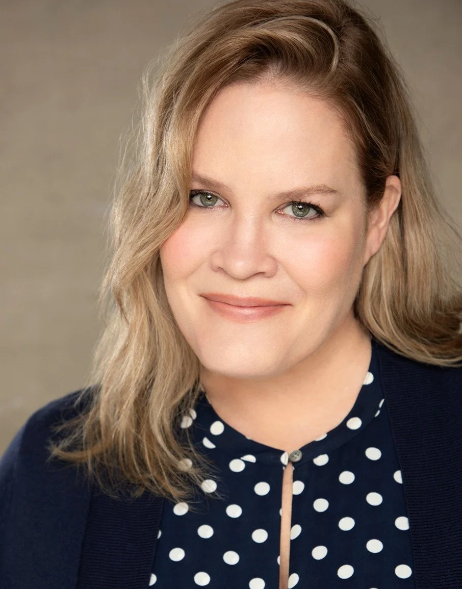 A woman with blonde hair wearing a navy blue polka dot blouse and a dark cardigan, smiling at the camera.