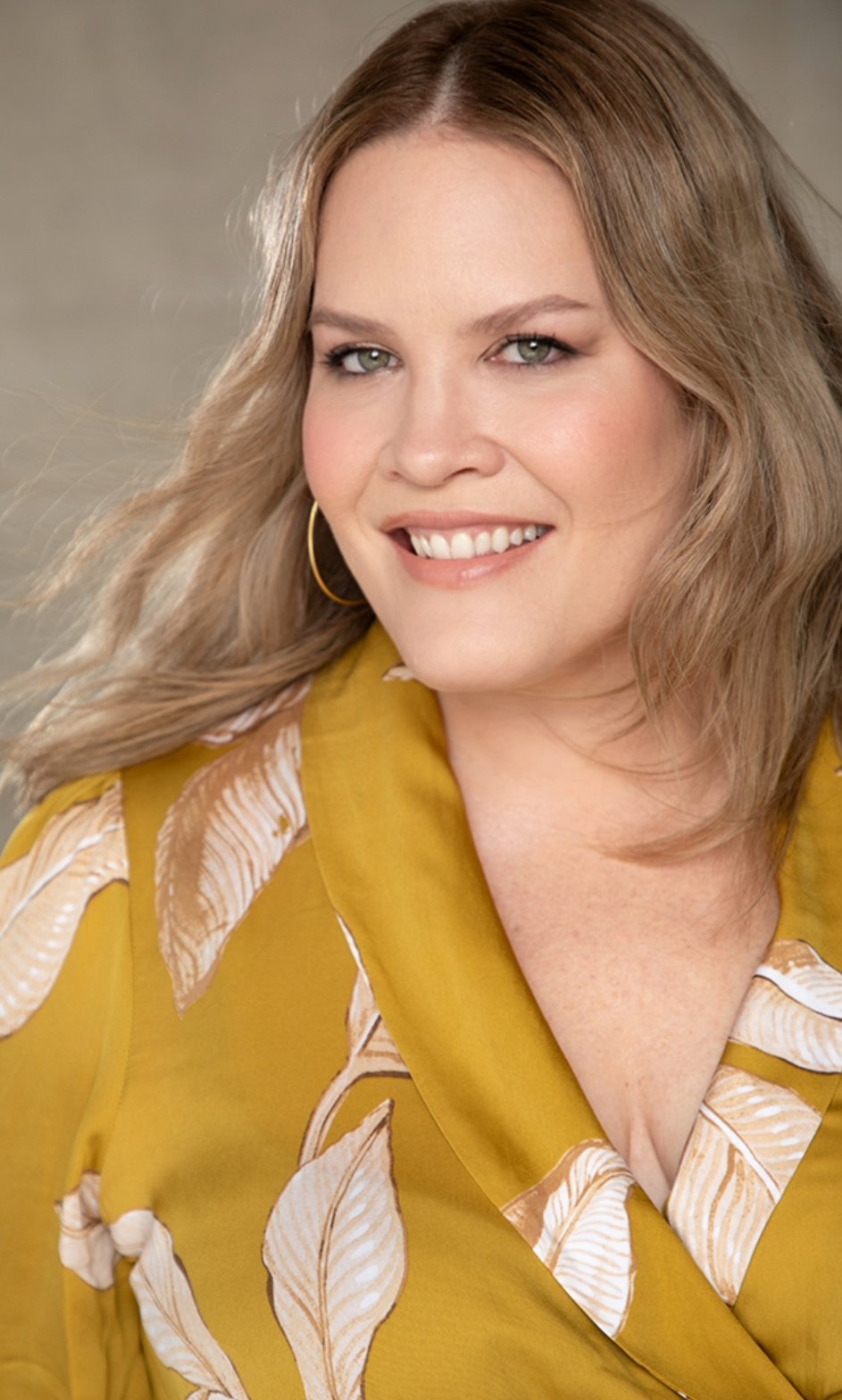 A woman with light brown hair, green eyes, and a friendly smile, wearing a yellow blouse with a leaf pattern and large hoop earrings.