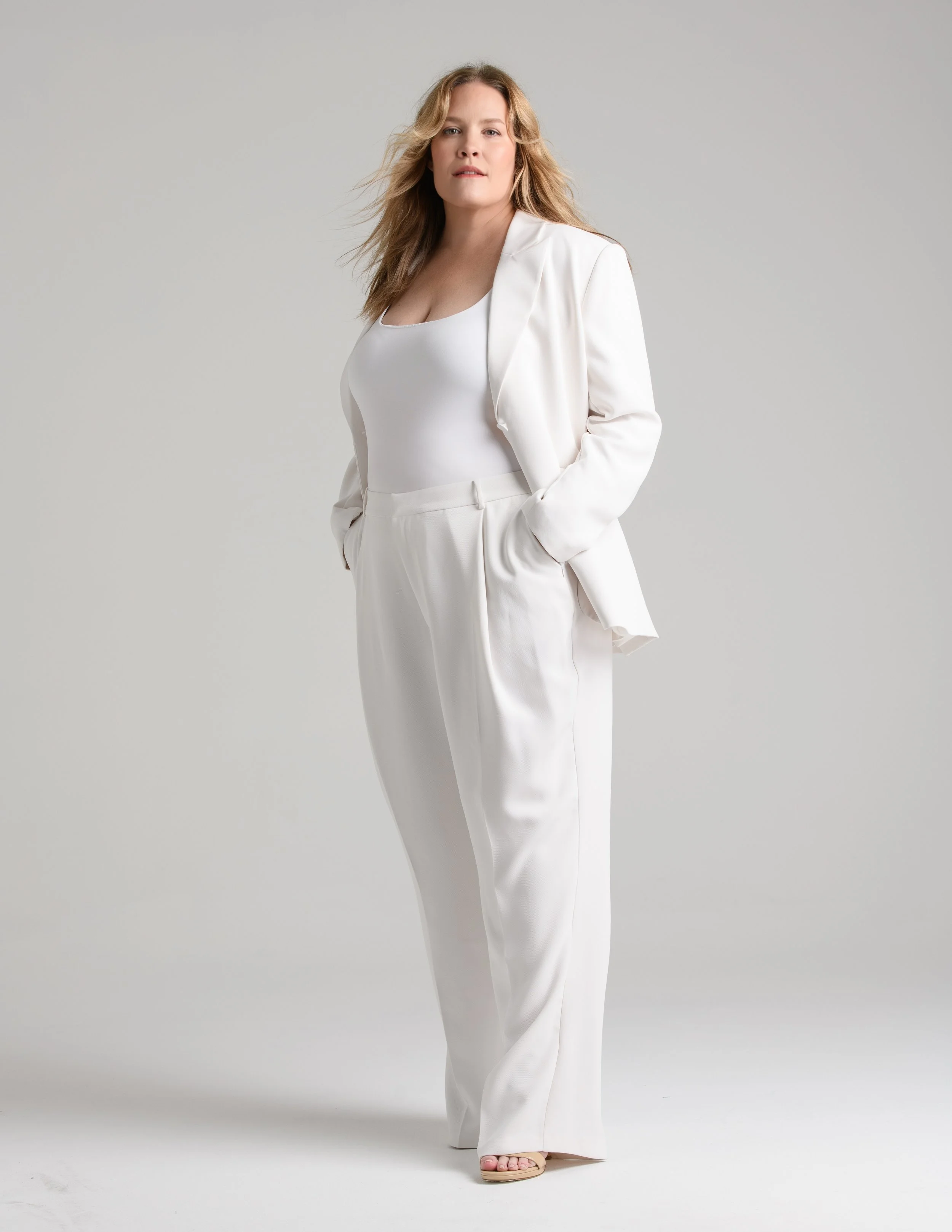 A woman with blonde hair wearing an all-white suit, pose with her hands in her pockets against a gray background.