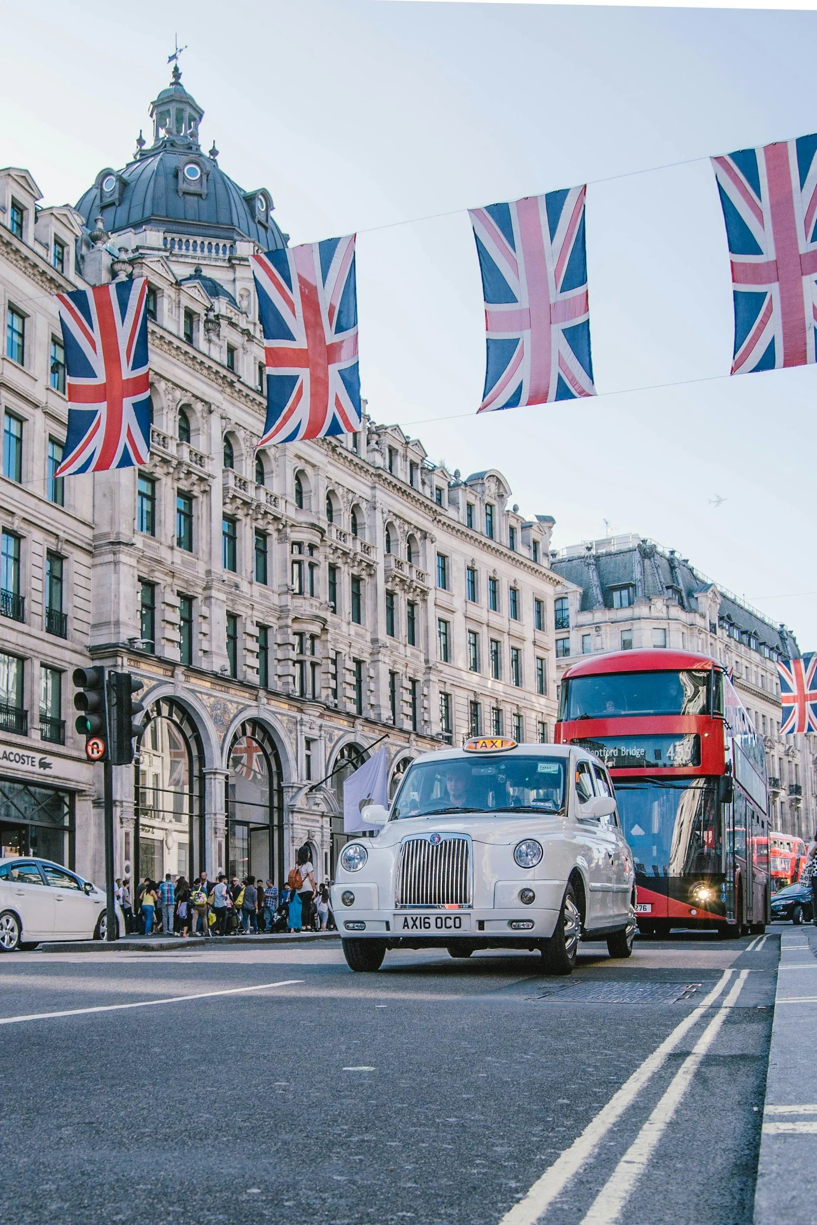 Street scene in London with Union Jack flags hanging above, a white taxi, red double-decker bus, and a crowd of pedestrians on the sidewalk in front of historic buildings.