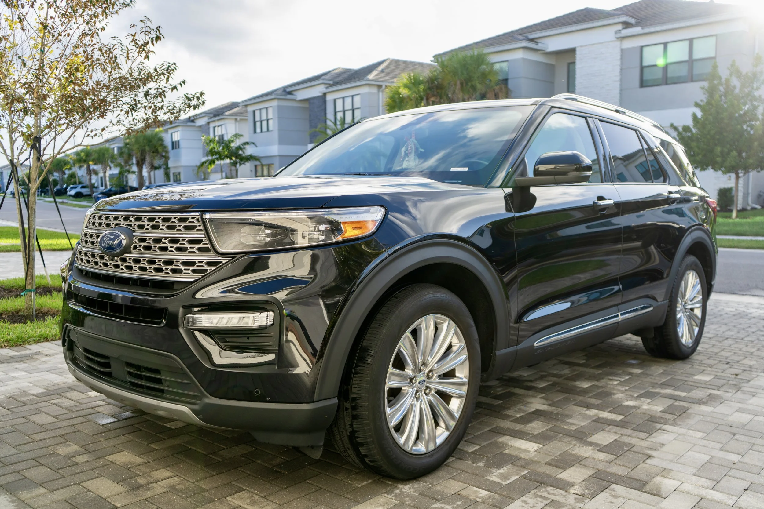 A black Ford SUV parked on a brick driveway in front of modern white residential buildings with palm trees and green lawns.
