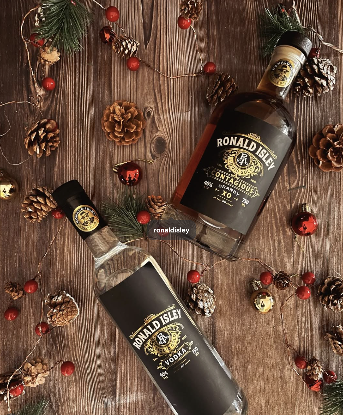 Two bottles of Ronald Isley liquor, one labeled vodka and the other brandy, lying on a wooden surface surrounded by pinecones, red berries, gold and red ornaments, and pine branches for holiday decoration.