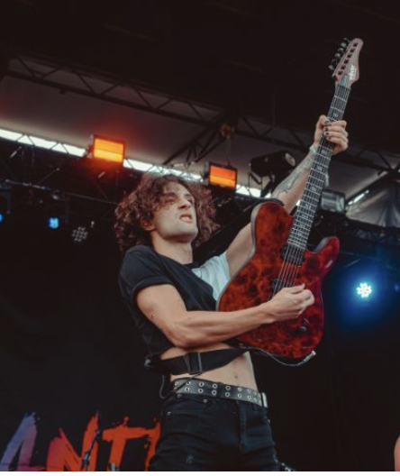 A female musician performing on stage, holding a red electric guitar, with stage lights overhead and a dark backdrop.