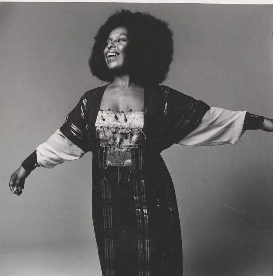 A woman with curly hair smiling and extending her arms outward in a black and white photo.
