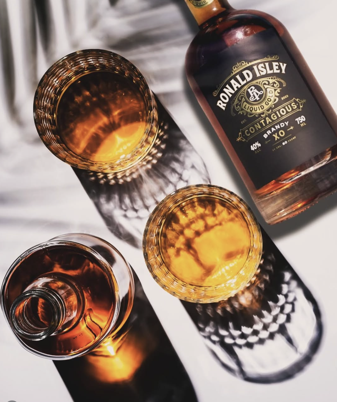 Bottle of Ronald Isley Contagious Brandy XO and three glasses with brandy, casting intricate shadow patterns on a white surface.