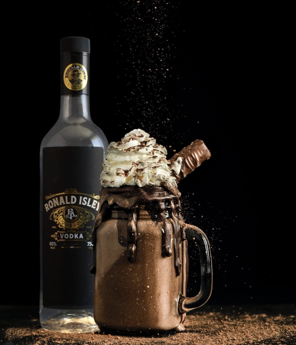 A chocolate milkshake in a mug topped with whipped cream and chocolate shavings, with a chocolate bar stuck into the whipped cream. Cocoa powder is sprinkled on top, with some falling from above. A bottle of Ronald Isle vodka stands behind the milkshake against a black background.