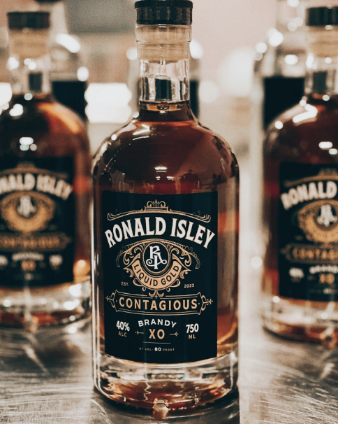 Close-up of a bottle of Ronald Isley Contagious Brandy with a black label, surrounded by other bottles on a wooden surface.