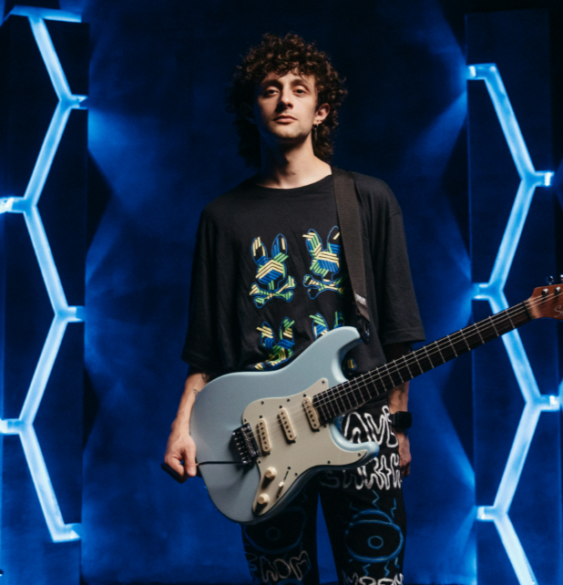 Young man holding a white electric guitar on a stage with blue hexagonal light designs in background.