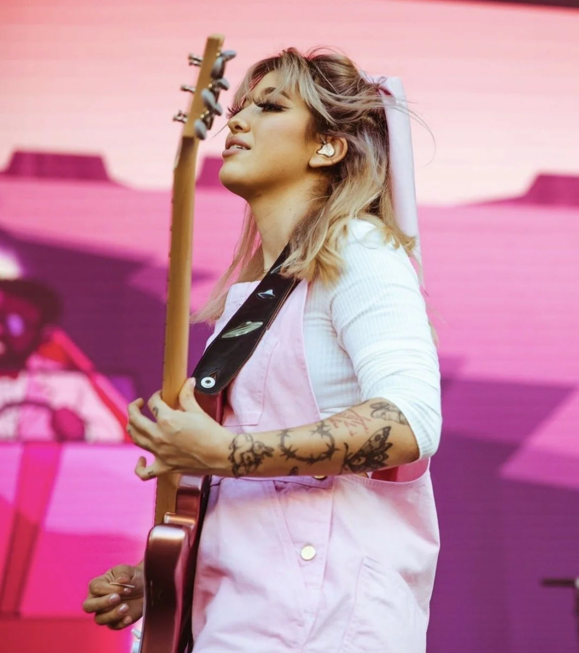 A woman with blonde hair, tattoos on her arms, and wearing a pink apron, playing an electric guitar on stage with a pink background.