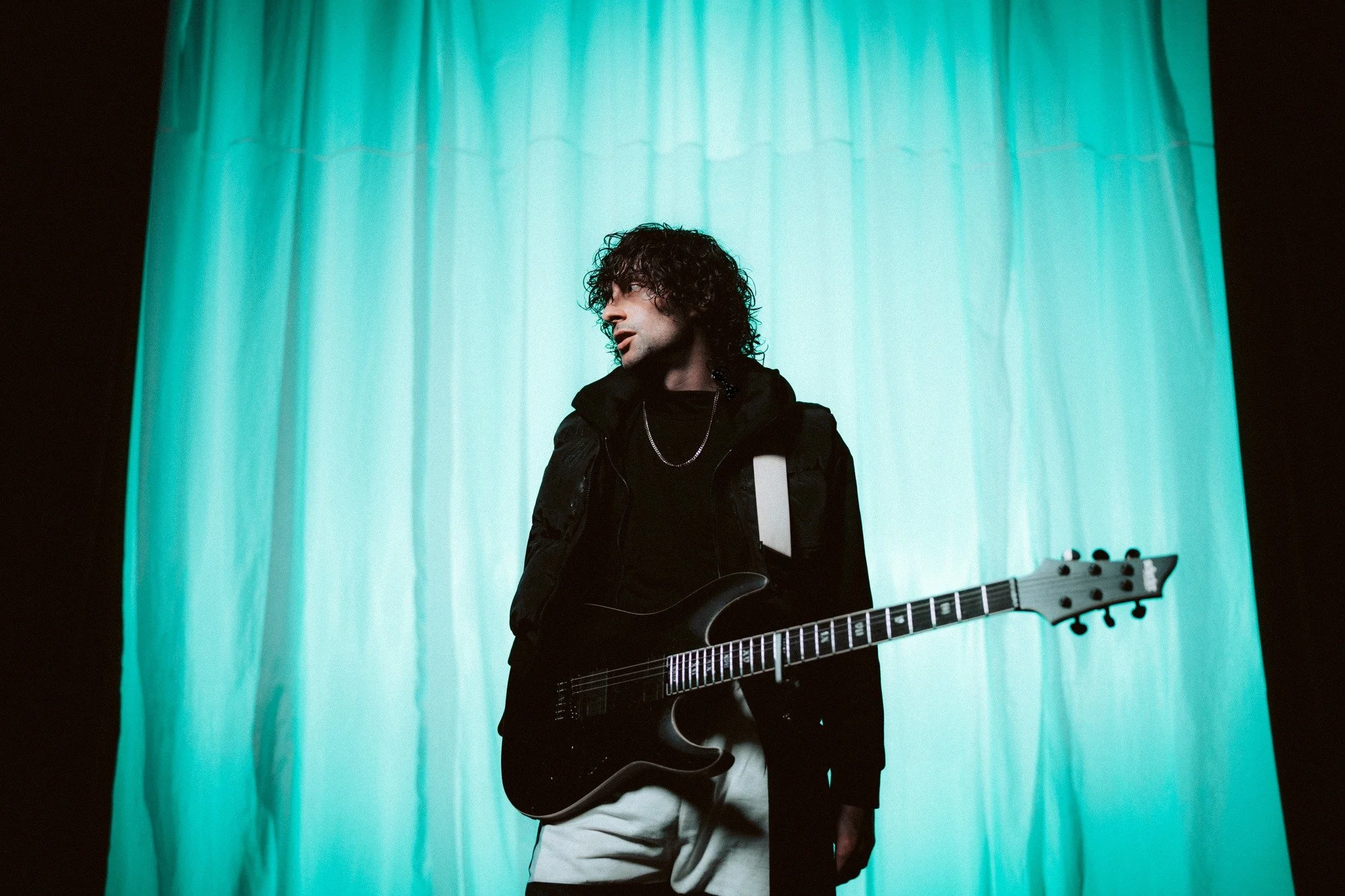 A young man with curly hair holding an electric guitar, standing in front of a teal curtain, wearing a black jacket and white pants.