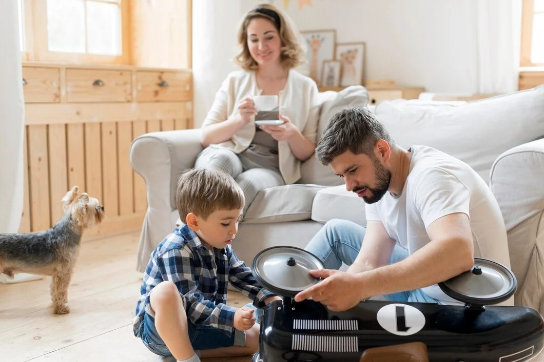 A family in a cozy living room with light wood furniture and framed pictures, including a woman sitting on a couch holding a cup, a man assembling a toy drone, and a young boy sitting on the floor playing with the drone. A small dog stands nearby looking at the family.